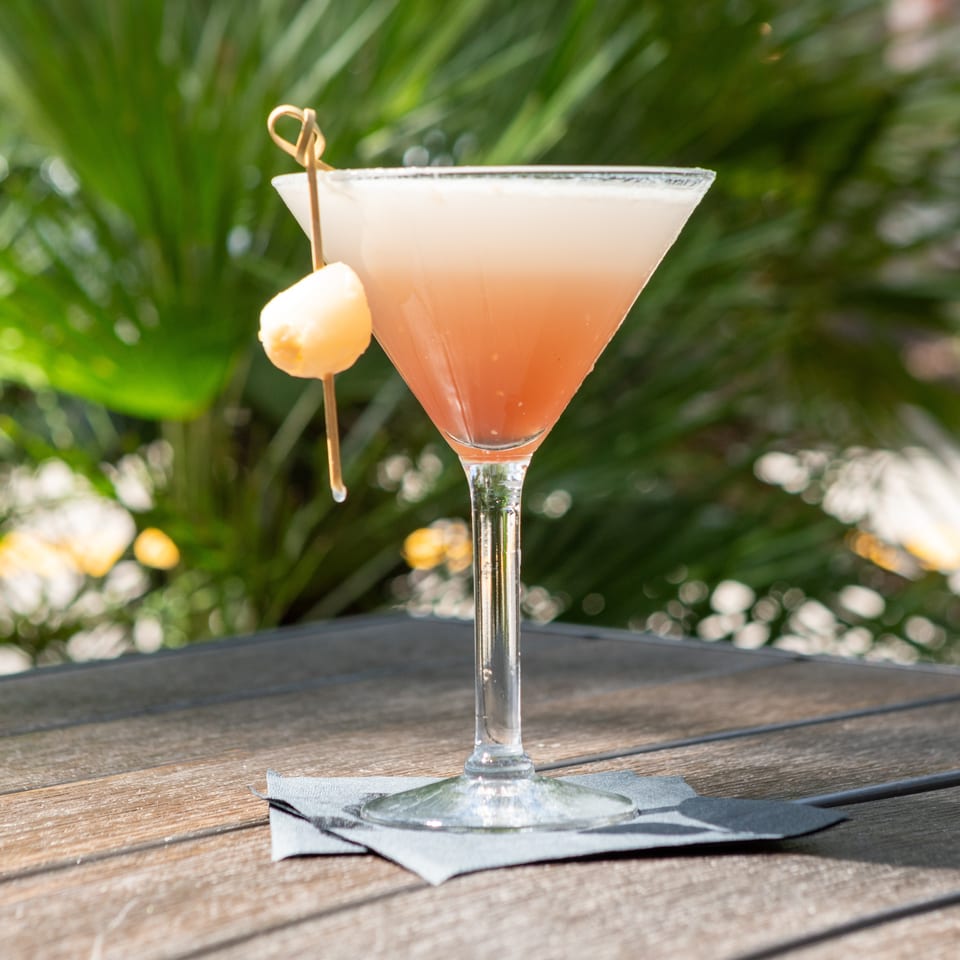 Pink cocktail in a sugar-rimmed martini glass, garnished with a fruit skewer, sits on a napkin-covered outdoor table.