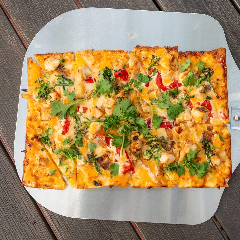 Rectangular cheesy pizza topped with chicken, peppers, broccoli, mushrooms, and fresh cilantro on a metal serving tray.