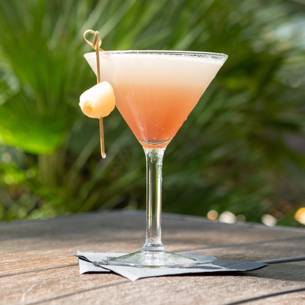 Peach-colored cocktail in a martini glass, garnished with a fruit skewer, sits on a napkin atop an outdoor wooden table.