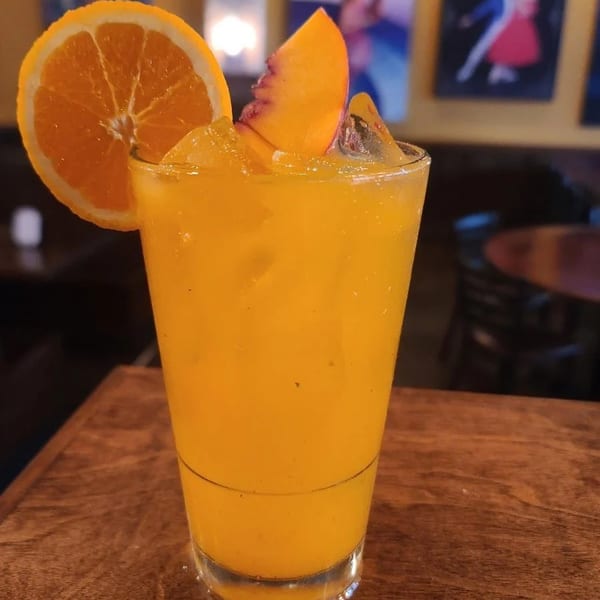 Orange cocktail with ice, garnished with an orange slice and peach wedge on a wooden table.