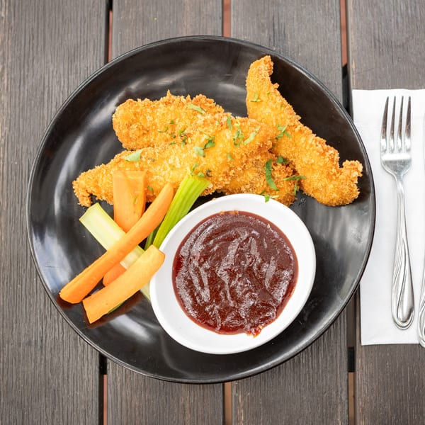 Breaded chicken tenders with carrot and celery sticks, barbecue dipping sauce, and parsley on a black plate.