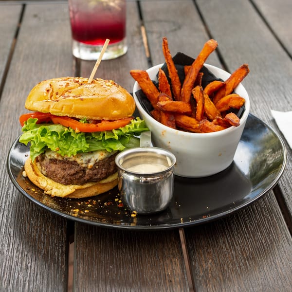 Cheeseburger with lettuce and tomato served alongside sweet potato fries and dipping sauce on a restaurant table.