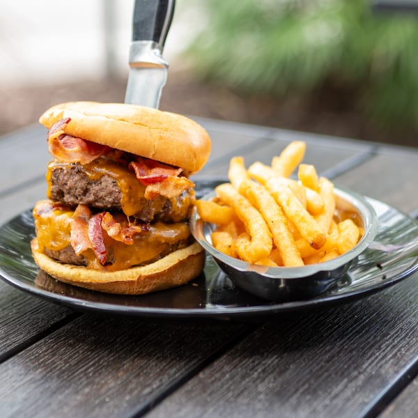Bacon cheeseburger with double beef patties and golden fries served on a black plate outdoors.