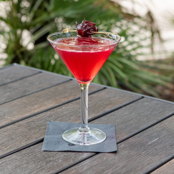 Bright red cocktail in a martini glass garnished with a dried flower, resting on a napkin atop an outdoor wooden table.