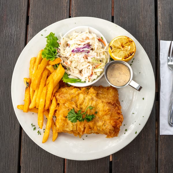 A plate of fried fish and fries with coleslaw, dipping sauce, grilled lemon, and parsley on a dark wooden table.