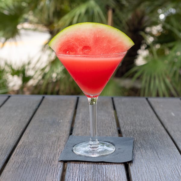 Bright red watermelon cocktail in a martini glass, garnished with a watermelon wedge, sits on a wooden outdoor table.
