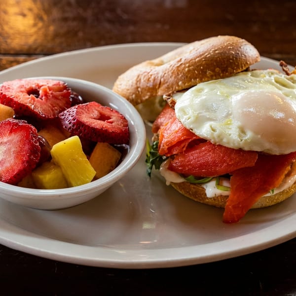 Bagel with smoked salmon, fried egg, and a side of fresh fruit on a white plate.