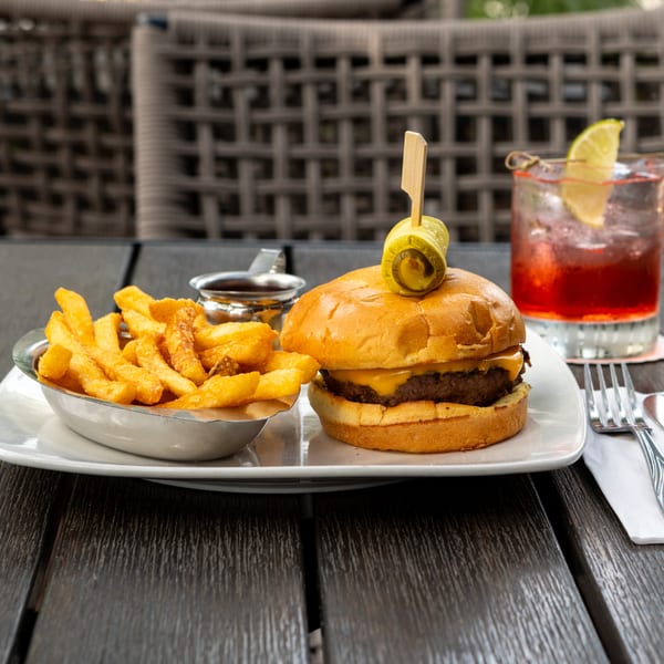 Cheeseburger with pickle garnish and fries on a plate, served beside a red citrus cocktail on an outdoor patio table.
