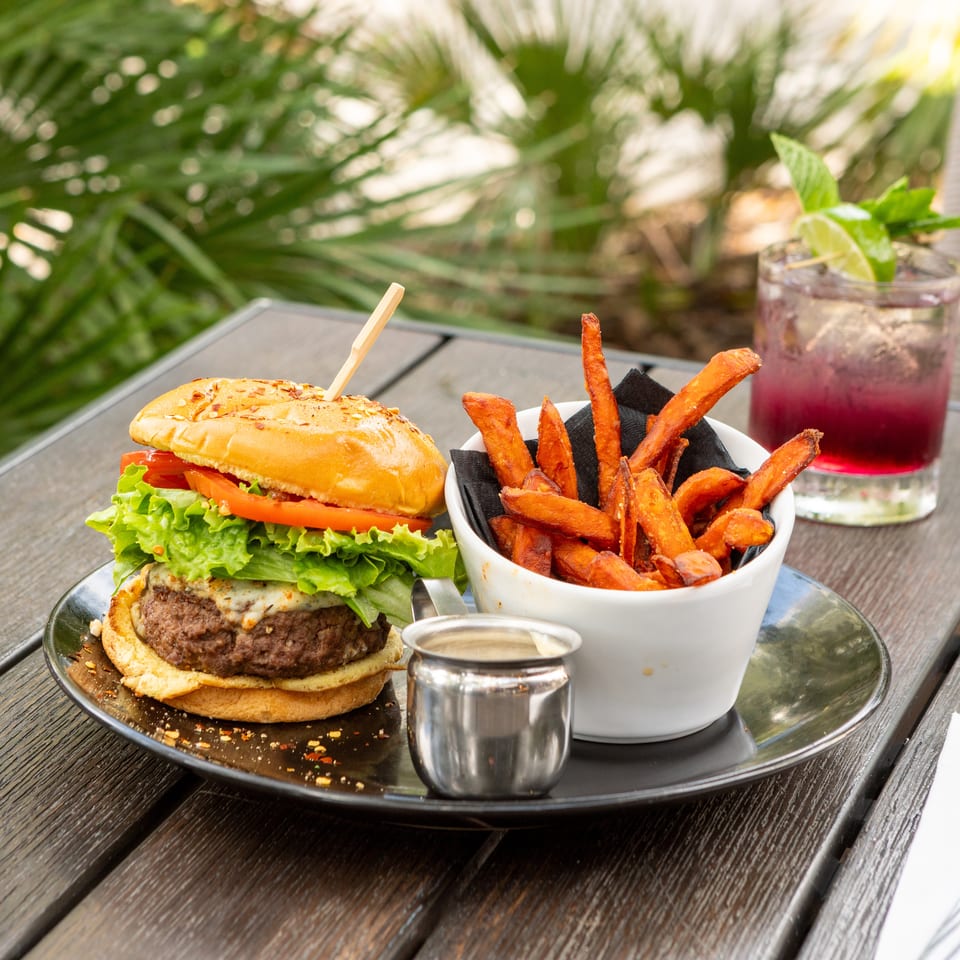 Gourmet burger with lettuce and tomato, sweet potato fries, dipping sauce, and a berry drink on an outdoor restaurant table.