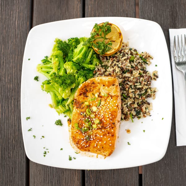 Grilled salmon fillet with sesame and scallions, steamed broccoli, mixed grains, lemon, and parsley on a white plate.