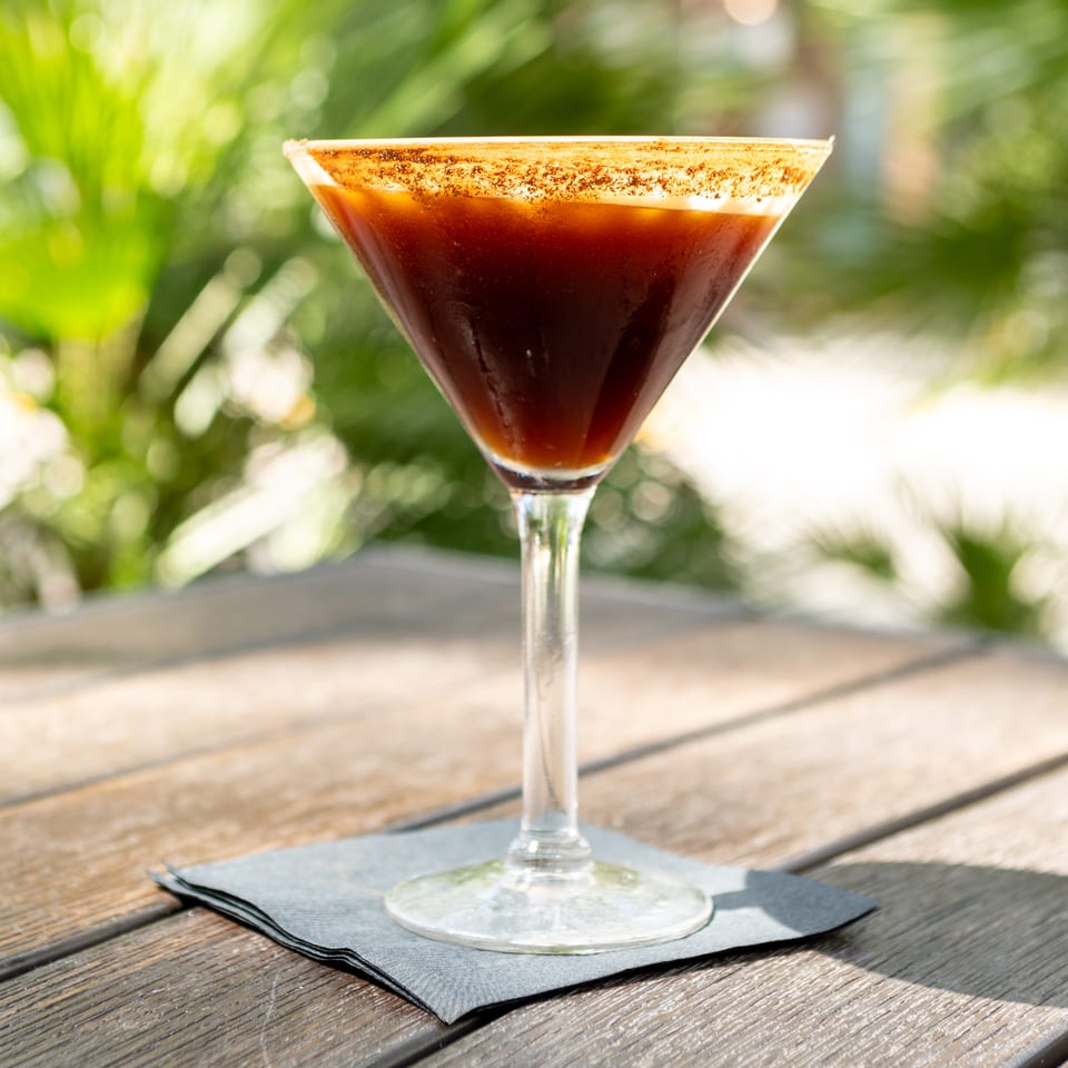 Dark cocktail in a sugar-rimmed martini glass on a black napkin atop an outdoor wooden table.