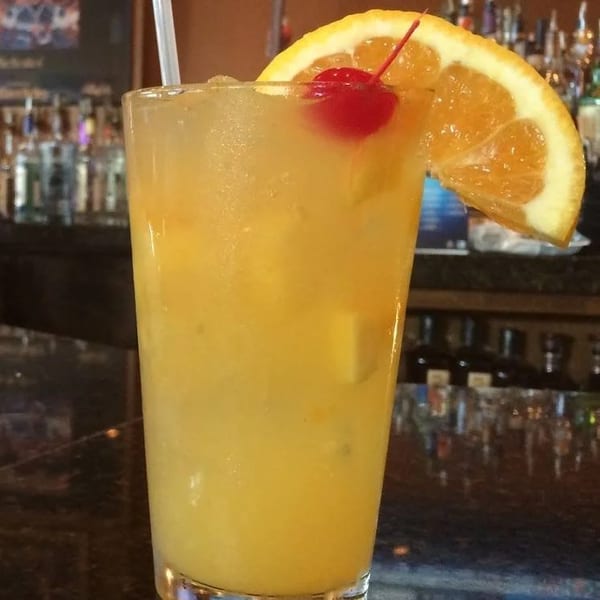 A tall glass of yellow cocktail with ice, an orange slice, and a cherry on a bar.