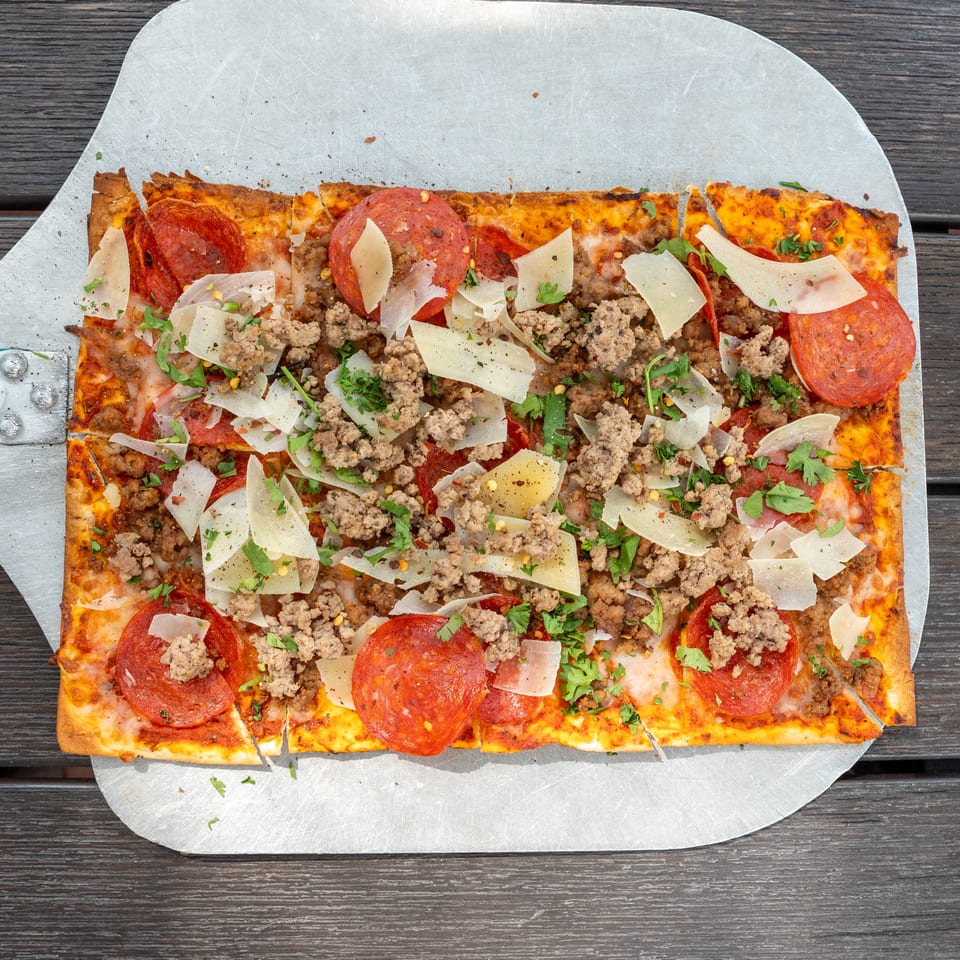 Rectangular pizza topped with pepperoni, ground meat, shaved cheese, herbs, and chili flakes on a metal pizza peel.